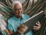 Senior man enjoys leisure time using a tablet in a colorful hammock.